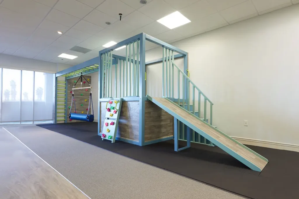 Sensory gym with rock climbing wall, slide, swing with beam, and monkey bars, featuring wooden finish and light blue beams