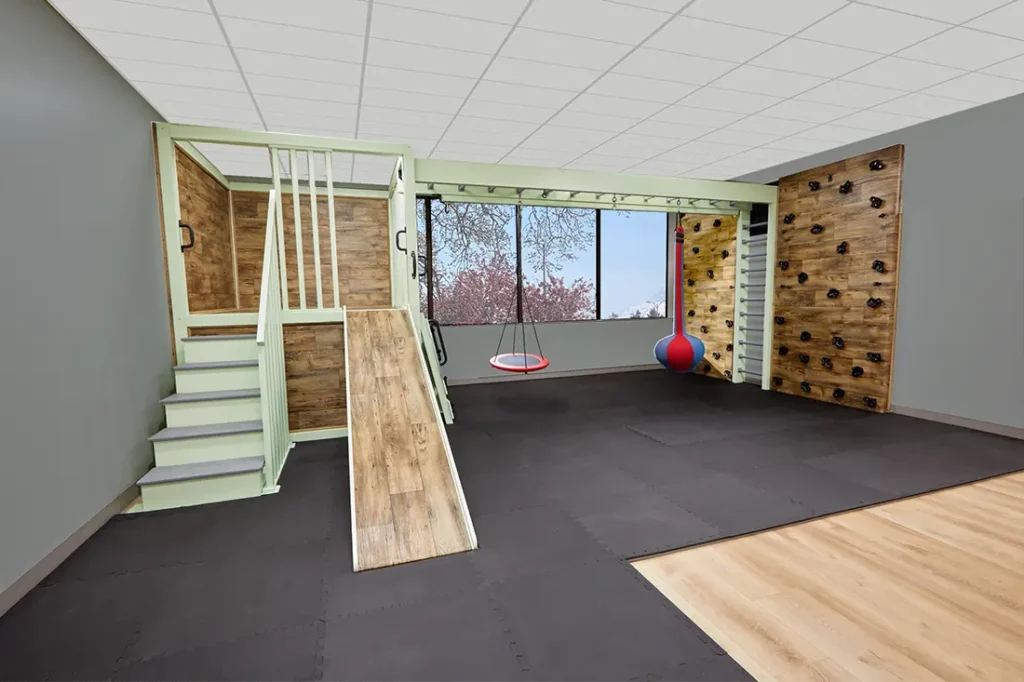 Large sensory gym with steps, slide, monkey bars, rock climbing walls, two swings including a ball swing, floor padding, and a window view