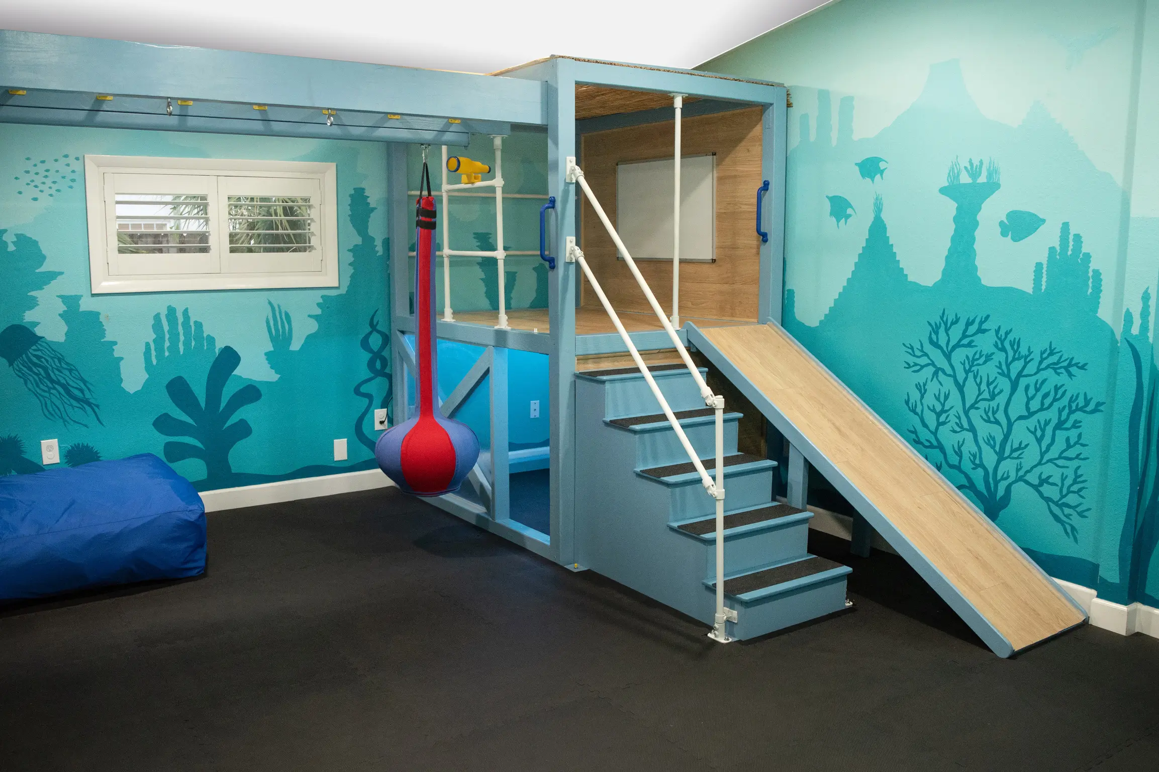 Sensory gym with steps, a slide, monkey bars, a ball swing, and a window view with underwater-themed walls