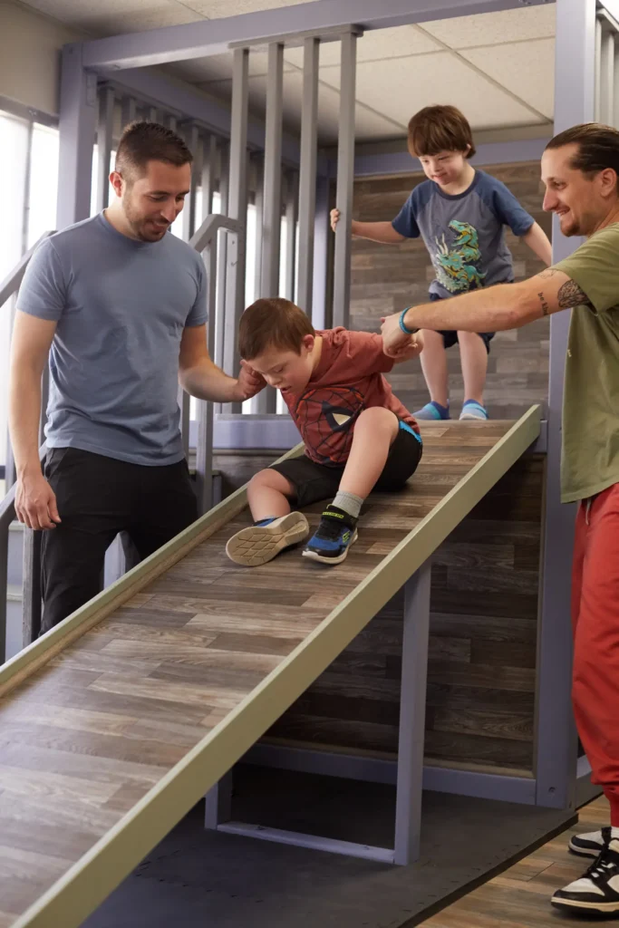 Children with special needs playing on a sensory gym slide with supportive adults.